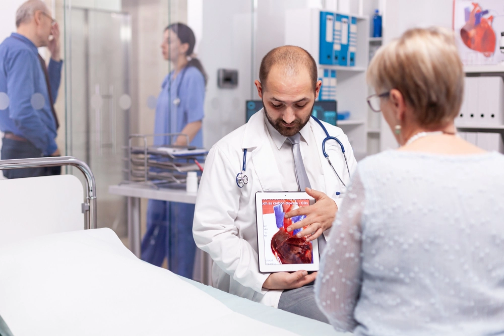 A specialist doctor using a digital tablet to explain a heart condition to a senior patient in a clinical setting.